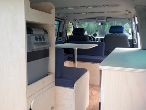 Aménagement de van en bois clair avec coin repas et glacière électrique.
